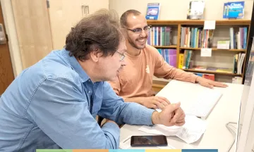 Two adults sitting at a workspace together looking at a computer 