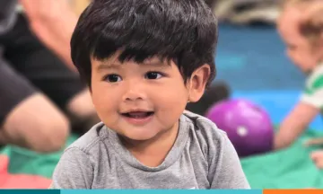 Smiling toddler in a gray shirt with another child and toys in the background.