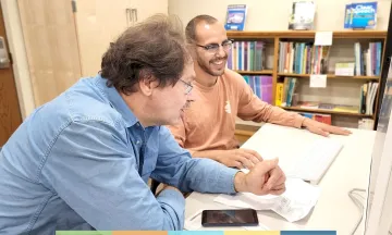 Two adults sitting at a workspace together looking at a computer 