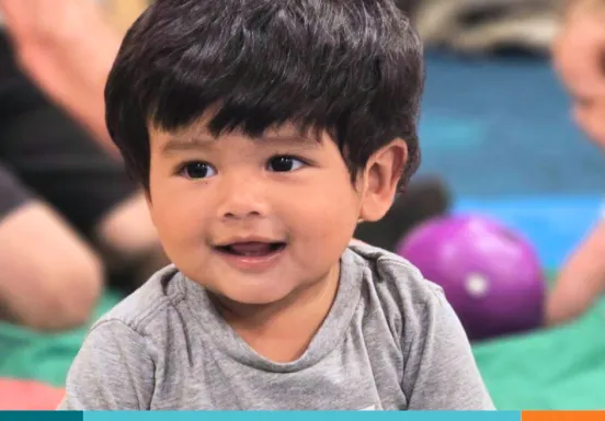 Smiling toddler in a gray shirt with another child and toys in the background.
