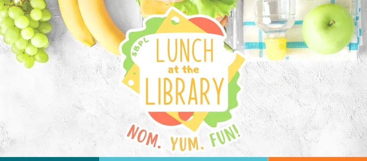 A colorful sandwich sits on a lunch table, with the text "Lunch at the Library" located on the bread and "Nom. Yum. Fun!" below.