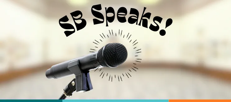 A microphone on a stand in the foreground with the text “SB Speaks!” above it on a transparent room background.