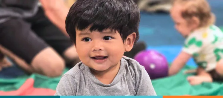 Smiling toddler in a gray shirt with another child and toys in the background.