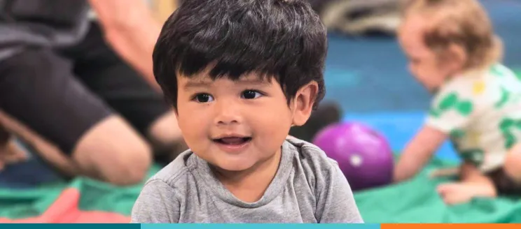 Smiling toddler in a gray shirt with another child and toys in the background.