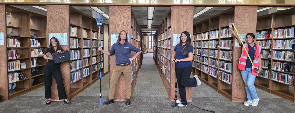4 people stand in the book stacks holding tools that can be found in the library's Tool Library collection